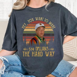 May include: Dark gray t-shirt featuring a retro design with a man wearing a cap and a colorful shirt. The text reads "HEY, YOU WANT TO MAKE $14 DPLARS THE HARD WAY" in a vintage font, set against a sunset background.