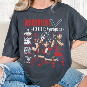 May include: Dark gray t-shirt featuring the text "RESIDENT EVIL X - CODE: Veronica-" in red and white. The graphic includes character illustrations in red and white, with a distressed vintage style.