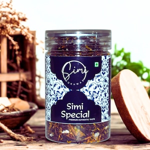May include: A clear jar filled with a dark mixture, labeled "Simi Special." The label is dark blue with white floral patterns and text. A wooden lid rests beside the jar. The background includes wooden crates and greenery, all on a wooden surface.