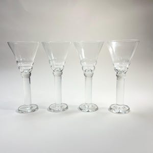 May include: Four clear glass stemmed glasses. Each glass has a conical bowl, a decorative stem with a bulbous design, and a circular base. The glasses are arranged in a row against a white background.