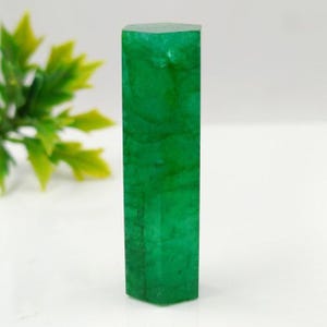 Könnte beinhalten: Ein lebendiger, smaragdgrüner Edelstein mit rechteckiger Form und facettierter Oberseite. Die polierte Oberfläche reflektiert das Licht und zeigt die natürlichen Muster und Farbvariationen des Steins. Der Edelstein wird vor einem weißen Hintergrund präsentiert.