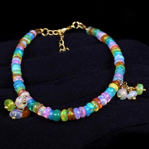 Puede incluir: Una pulsera de cuentas con pequeñas piedras redondas e iridiscentes en tonos multicolor, incluyendo azul, verde, naranja y rosa. La pulsera incluye una cadena extensora de metal dorado y dos pequeños grupos de piedras colgantes en los extremos.