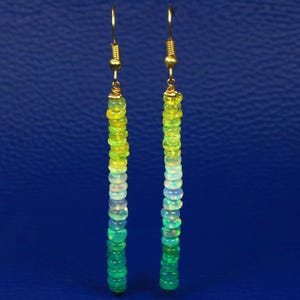 Könnte beinhalten: Ein Paar Ohrhänger mit einem Farbverlauf aus grünen und gelben Opalperlen. Die Perlen sind aufgereiht und an goldfarbenen Ohrhaken befestigt. Das Design ist vertikal, vor blauem Hintergrund. Die Ohrringe sind ca. 7 cm lang.