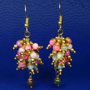 Könnte beinhalten: Ein Paar handgefertigte Ohrringe mit Clustern aus rosa, weißen und grünen Perlen. Die Perlen sind mit goldenem Draht umwickelt und an goldfarbenen Ohrhaken aufgehängt. Eine kleine, dunkle Perle baumelt von der Unterseite jedes Ohrrings.