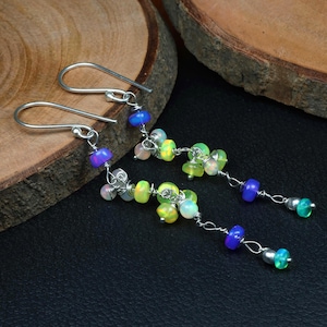 Könnte beinhalten: Ohrringe mit bunten Perlen in Blau, Grün, Gelb und Rosa. Die Perlen sind auf Silberdraht aufgefädelt und an silbernen Ohrhaken befestigt. Die Ohrringe werden auf einer dunklen Oberfläche präsentiert.