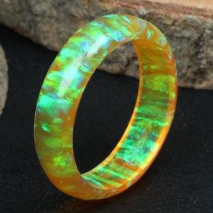 Könnte beinhalten: Ein lebendiger, schillernder Ring mit einem faszinierenden Farbenspiel. Der Ring zeigt eine Mischung aus Orange-, Grün- und Blautönen, die einen fesselnden visuellen Effekt erzeugt. Das Design des Rings ist schlicht, aber die Farbvariationen machen ihn besonders.