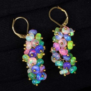 May include: A pair of dangle earrings featuring clusters of small, round, iridescent opal beads in shades of pink, blue, and green. Each bead is accented with tiny gold-toned metal spheres, and the earrings are attached to gold-toned lever-back hooks.