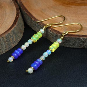 Könnte beinhalten: Ein Paar handgefertigte Ohrringe mit goldfarbenen Ohrhaken. Die Ohrringe weisen einen Farbverlauf aus bunten Perlen auf, der von Blau nach Grün übergeht, mit weißen Opalperlen dazwischen. Die Ohrringe werden auf einer dunklen Oberfläche präsentiert.