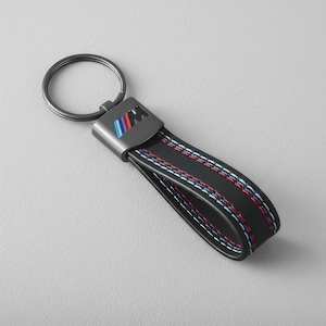 May include: Black leather keychain with a metal ring and a rectangular metal plate featuring the BMW M logo in blue, red, and light blue. The keychain strap has white, red, and blue stitching.