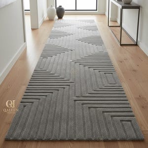 Op de afbeelding: Een lang, rechthoekig grijs tapijt met een geometrisch patroon van vierkanten en lijnen. Het tapijt ligt op een lichtgekleurde houten vloer. Het tapijt is een gangloper.