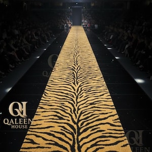 Puede incluir: Una larga alfombra rectangular con un estampado de rayas de tigre en dorado y negro. La alfombra es el punto focal de una pasarela de moda, con un suelo negro y asientos para el público a ambos lados. La marca "QALEEN HOUSE" es visible.