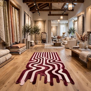 Puede incluir: Una alfombra rectangular con un patrón ondulado similar a un líquido en color burdeos intenso y rosa claro. La alfombra está colocada sobre un suelo de madera clara en una sala de exposición llena de rollos textiles y decoración.