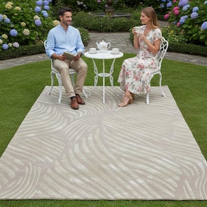 May include: A beige outdoor rug with a wave pattern is laid on green grass. Two people are seated at a small white table, enjoying tea. The rug is rectangular and has a textured design. The scene is set in a garden with colorful flowers.