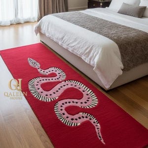 Puede incluir: Una alfombra rectangular roja con un diseño de serpiente rosa y blanca. La alfombra está colocada frente a una cama con un edredón blanco y un camino de cama marrón. La alfombra tiene el texto "QALEEN HOUSE".
