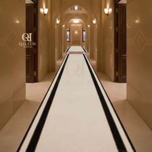 Op de afbeelding: Een binnenhal met een crèmekleurig loperkleed, begrensd door zwarte strepen. De hal heeft gewelfde deuropeningen, wandkandelaars en het "QALEEN HOUSE" logo.
