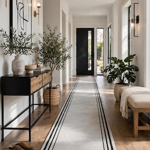 May include: A long rectangular runner rug featuring a light gray center with double black border stripes. The rug is positioned in a hallway with a black console table, a wooden bench, potted plants, and a black door visible in the background.