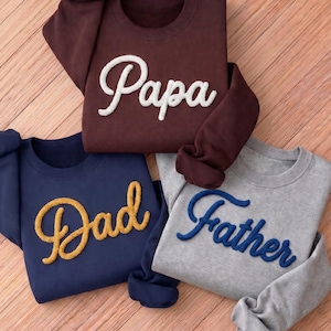 May include: Three crewneck sweatshirts in burgundy, navy blue, and gray. Each sweatshirt features a script word: "Papa" in white, "Dad" in yellow, and "Father" in blue. The sweatshirts are displayed on a wooden surface.