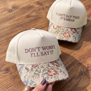May include: Two cream-coloured baseball caps with floral tapestry brims. The front cap features the embroidered text "DON'T WORRY, I'LL SAY IT" in brown, while the back cap features the embroidered text "I CAN'T SAY THAT, IT'S MEAN" in brown.