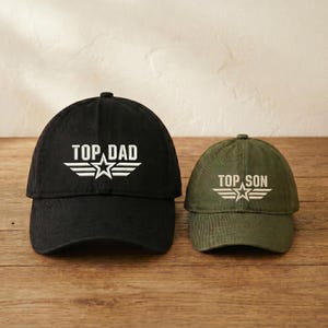 May include: Two baseball caps, one black and one olive green, sit on a wooden surface. The black cap reads "TOP DAD" in white block letters above a white star and wing design. The olive green cap reads "TOP SON" in the same style.