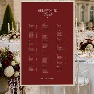 Könnte beinhalten: Ein rechteckiges Hochzeitssitzplan-Schild mit tiefrotem Hintergrund und weißem Text. Das Schild trägt die Überschrift "OUR FAVORITE People" und listet die Namen der Gäste alphabetisch sortiert auf, endend mit "STACEY & GREGORY".
