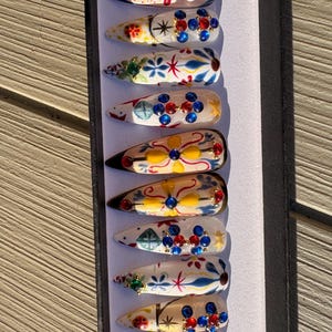 Peut inclure: Un ensemble de neuf faux ongles longs en forme d'amande. Chaque ongle est peint avec un motif floral unique en rouge, bleu, jaune et vert, orné de strass et d'ornements dorés. Les ongles sont présentés dans une boîte noire.