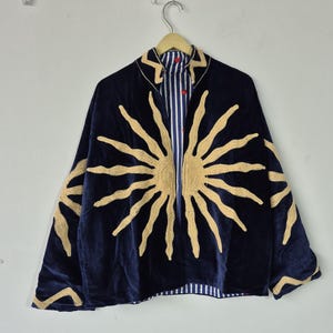Puede incluir: Una chaqueta de terciopelo azul oscuro con un gran diseño de sol bordado en dorado en la parte delantera y las mangas. La chaqueta tiene un cuello alto y una abertura frontal que revela un forro de rayas verticales blancas y azules con pequeños corazones rojos.