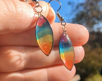 Könnte beinhalten: Ein Paar handgefertigte, tropfenförmige Ohrringe. Die Ohrringe weisen einen lebendigen Regenbogenverlauf aus Farben auf, darunter Rot, Orange, Gelb, Grün und Blau. Die Ohrringe sind an silberfarbenen Haken befestigt.