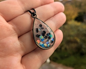 Puede incluir: Colgante en forma de lágrima con un centro de resina transparente lleno de purpurina negra e iridiscente. El colgante tiene un marco de metal color bronce y cuelga de un cordón negro. La purpurina refleja tonos azules, verdes y naranjas.
