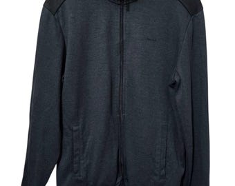 May include: A dark gray zip-up jacket with black shoulder accents and a stand-up collar. The jacket has a full-length zipper, two front pockets, and the Calvin Klein logo embroidered on the chest. The jacket is hanging on a black hanger.