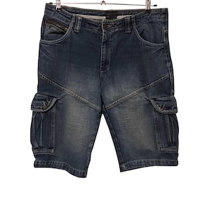 Puede incluir: Pantalones cortos vaqueros azules con múltiples bolsillos cargo y un detalle de cremallera en el bolsillo delantero. Los pantalones cortos cuelgan de una percha metálica sobre un fondo blanco. Los pantalones cortos tienen un lavado descolorido.