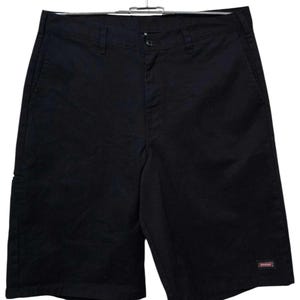 Könnte beinhalten: Schwarze Shorts hängen an einem Metallbügel. Die Shorts haben einen Knopfverschluss und Taschen. Ein kleines rot-weißes Logo ist auf dem rechten Bein sichtbar. Die Shorts sind aus robustem Material gefertigt.