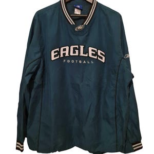 Peut inclure: Veste de football des Eagles de couleur sarcelle avec le nom de l'équipe en lettres blanches. La veste a un col en V avec des rayures noires et blanches, et le logo des Eagles est sur la poitrine. Les manches ont des poignets rayés.