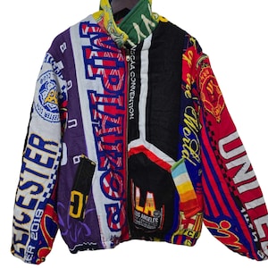 Op de afbeelding: Een kleurrijke patchworkjas met verschillende patronen en tekst. De jas heeft een mix van kleuren, waaronder rood, blauw, paars en zwart. De tekst bevat "Leicester City", "Los Angeles" en "NSGAA Convention".