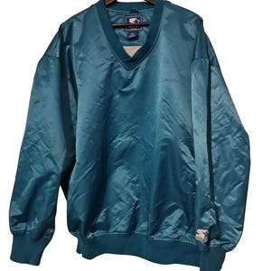 Könnte beinhalten: Türkisfarbene Vintage Starter Windjacke mit V-Ausschnitt und elastischen Bündchen und Saum. Die Jacke hängt an einem Holzbügel. Das Starter-Logo ist auf dem Ärmel zu sehen.