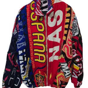 Może przedstawiać: Kurtka zapinana na zamek z żywym patchworkowym wzorem. Kurtka zawiera słowo "ESPANA" w dużych żółtych literach na czerwonym tle. Dodatkowy tekst i grafika w różnych kolorach zdobią kurtkę, w tym frazę "Orgulloso Patrocinador de los Campeones del Mundo."