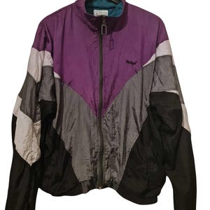 Könnte beinhalten: Vintage Trainingsjacke im Colorblock-Design. Die Jacke hat eine lila obere Brust, mit grauen, schwarzen und weißen Einsätzen. Sie hat einen durchgehenden Reißverschluss, einen Stehkragen und ein kleines Logo auf der Brust. Die Jacke hängt an einem schwarzen Kleiderbügel.