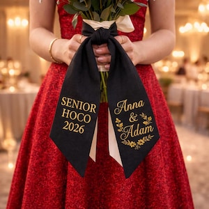 Könnte beinhalten: Schwarze Schärpe mit goldenem Text „SENIOR HOCO 2026“ und „Anna & Adam“ mit floralen Akzenten. Die Schärpe ist um einen Blumenstrauß gebunden, der von einer Person in einem roten Kleid gehalten wird.