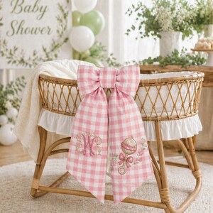 May include: A wicker bassinet with a pink and white gingham bow. The bow is embroidered with a floral monogram and a rattle design. A white blanket is inside the bassinet. The background includes a sign that says "Baby Shower".