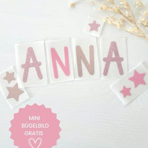 Könnte beinhalten: Bügelbilder mit dem Namen ANNA in rosa Glitzer- und einfarbigen rosa Buchstaben auf einzelnen transparenten Trägerfolien, ergänzt durch mehrere kleine sternförmige Bügelbilder in rosa Glitzer und Rosa sowie ein rosa kreisförmiges Etikett mit Text.