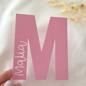 Könnte beinhalten: Ein rosa Vinyl-Aufkleber mit einem großen Großbuchstaben M, wobei der Name Malia in einer weißen Schreibschrift vertikal an der linken Seite des Buchstabens steht. Der Aufkleber befindet sich auf einer transparenten Klebefolie.