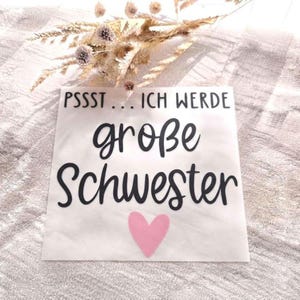 May include: A white square decal featuring the black text PSSST... ICH WERDE große Schwester and a small pink heart graphic. The decal is displayed with a decorative arrangement of dried beige and brown grasses positioned above it.
