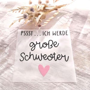 May include: A square white sign featuring the black text PSSST... ICH WERDE große Schwester with a small pink heart icon below the lettering. The sign is decorated with a cluster of dried beige and light purple flowers at the top edge.