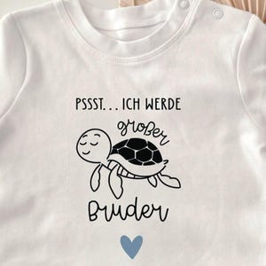 Pode incluir: Body de bebé branco com um gráfico preto de uma tartaruga e o texto alemão "PSSST... ICH WERDE größer Bruder" e um coração azul. O body tem fechos de pressão no ombro.