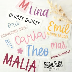 May include: A collection of adhesive vinyl decals featuring various names and phrases including Lina, Grober Bruder, Emil, Schulkind 2026, Carla, Kleiner Bruder, Mali, Theo, Malia, Noah Est 2025, and Kleine Schwester in diverse fonts, colors, and sizes.