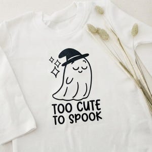 Könnte beinhalten: Ein weißes Langarmshirt mit einer schwarzen Grafik eines lächelnden Geistes, der einen Hexenhut trägt, sowie drei kleinen Sternen. Unter dem Geist steht der Text "TOO CUTE TO SPOOK" in einer fetten, schwarzen, abgerundeten Schrift.