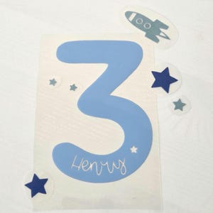 May include: A light blue number three decal featuring the name Henry written in white script. The design includes several dark blue and light blue star-shaped accents and a small rocket ship graphic, all arranged on a clear adhesive backing sheet.