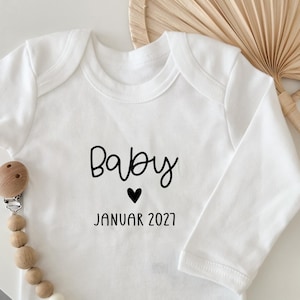 Könnte beinhalten: Weißer Baby-Body mit langen Ärmeln. Der Text "Baby" ist in handschriftlicher Form gedruckt, darunter ein kleines schwarzes Herz, gefolgt von "JANUAR 2027". Links befindet sich ein Schnullerhalter aus Holz mit Holzperlen.