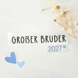 以下が含まれることがあります： 黒い文字でGROBER BRUDER、水色の文字で2027、そしてサイズの異なる3つの水色のハートが描かれたビニールステッカーセット。右上にはベージュのドライフラワーの小枝が添えられています。