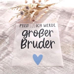 May include: A square white decal featuring the black text PSSST... ICH WERDE großer Bruder and a light blue heart icon below the text. The decal is displayed on a textured surface with a cluster of dried beige and brown grasses positioned above it.