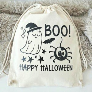 Könnte beinhalten: Ein cremefarbener Kordelzug-Rucksack aus Segeltuch mit einem schwarzen Grafikdruck eines Geistes mit Hut, einer Spinne, Sternen und einer Fledermaus. Der Text lautet BOO! und HAPPY HALLOWEEN in fetter schwarzer Schrift. Mit passenden Kordeln.